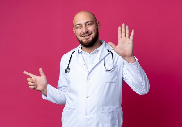 Pleased young bald male doctor wearing medical robe and stethoscope showing different numbers isolated on pink wall