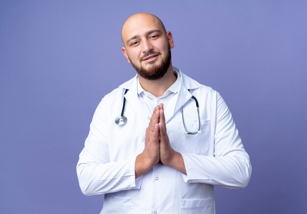 Pleased young bald male doctor wearing medical robe and stethoscope showing pray gesture isolated on blue background