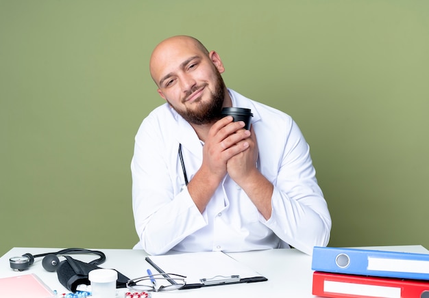 Pleased young bald male doctor wearing medical robe and stethoscope sitting at desk work with medical tools holding cup of coffee isolated on green background