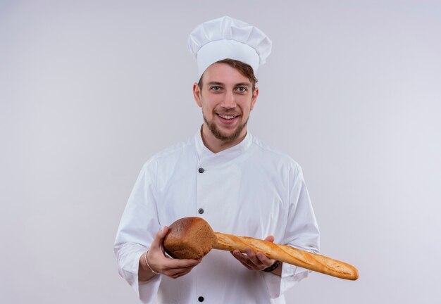 A pleased young bearded chef man wearing white cooker uniform and hat holding baguette bread with loaf bread while looking on a white wall