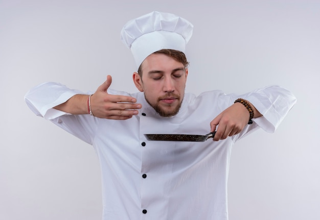 Free photo a pleased young bearded chef man wearing white cooker uniform and hat smelling frying pan on a white wall