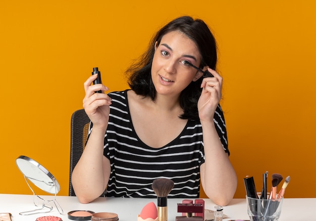 Free photo pleased  young beautiful girl sits at table with makeup tools applying mascara isolated on orange wall