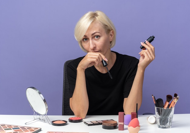 Free photo pleased young beautiful girl sits at table with makeup tools holding mascara isolated on blue wall