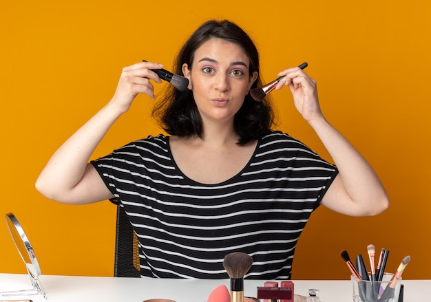 Free photo pleased young beautiful girl sits at table with makeup tools holding powder brushes around face isolated on orange wall