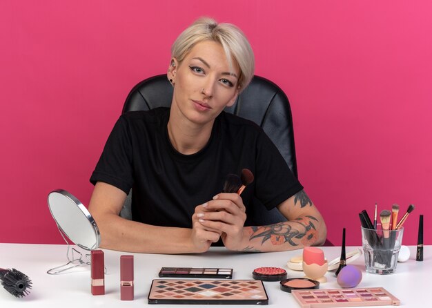 Pleased  young beautiful girl sits at table with makeup tools holding powder brushes isolated on pink wall