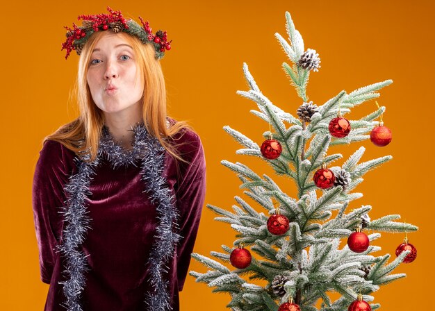 Pleased young beautiful girl standing nearby christmas tree wearing red dress and wreath with garland on neck showing kiss gesture isolated on orange background