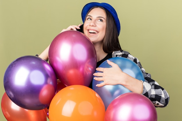 Pleased young beautiful girl wearing blue hat standing behind balloons speaks on phone 