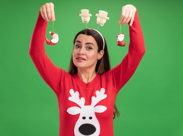 Pleased young beautiful girl wearing christmas sweater with christmas hair hoop raising toys isolated on green background