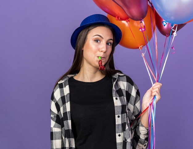 Pleased young beautiful girl wearing party hat holding balloons blowing party whistle 