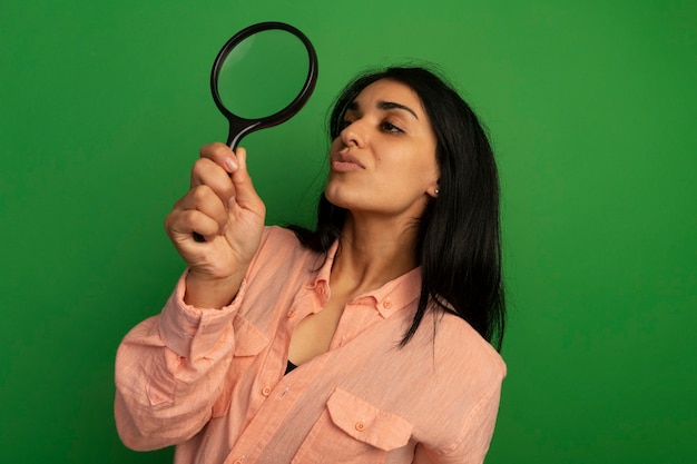 Pleased young beautiful girl wearing pink t-shirt raising and looking at magnifier isolated on green wall