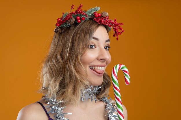 Pleased young beautiful girl wearing purple dress with wreath with garland on neck holding christmas candy isolated on brown background