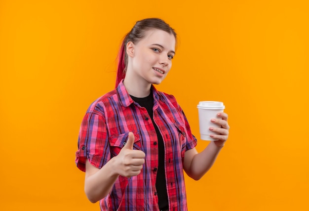 Pleased young beautiful girl wearing red shirt holding cup of coffee her thumb up on isolated yellow background with copy space