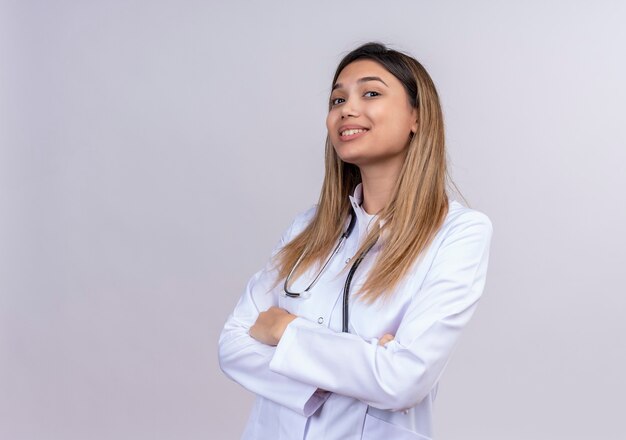 Pleased young beautiful woman doctor wearing white coat with stethoscope looking confident standing with arms crossed