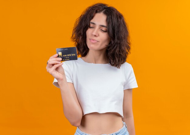 Pleased young beautiful woman holding card and looking at it on isolated orange wall with copy space