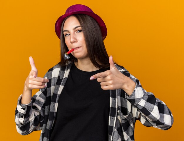 Pleased young beautiful woman wearing party hat blowing party whistle points at front