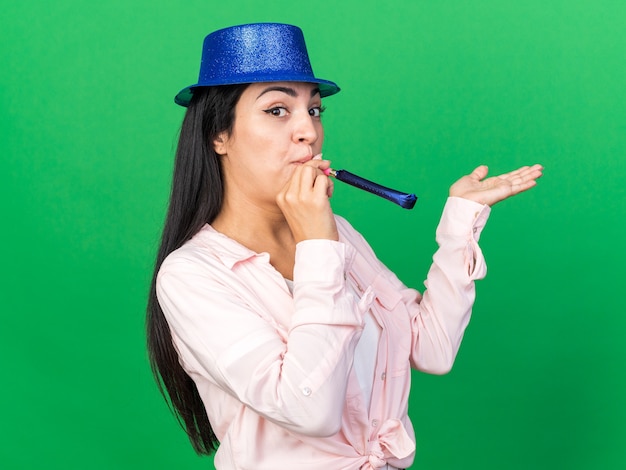 Free photo pleased young beautiful woman wearing party hat blowing party whistle spreading hand isolated on green wall