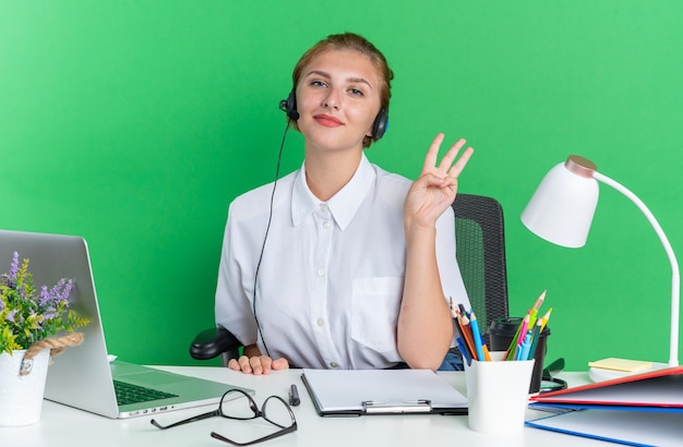 Free photo pleased young blonde call centre girl wearing headset sitting at desk with work tools showing three with hand