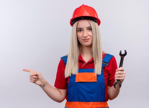 Pleased young blonde engineer builder girl in uniform holding open end wrench pointing at left side on isolated white space