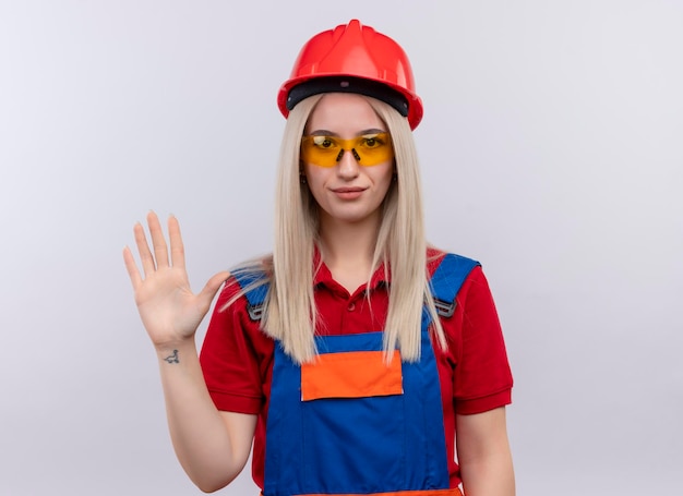 Pleased young blonde engineer builder girl in uniform wearing safety glasses gesturing hi on isolated white space