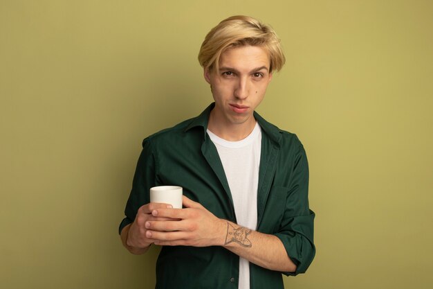 Pleased young blonde guy wearing green t-shirt holding cup of tea crossing hands