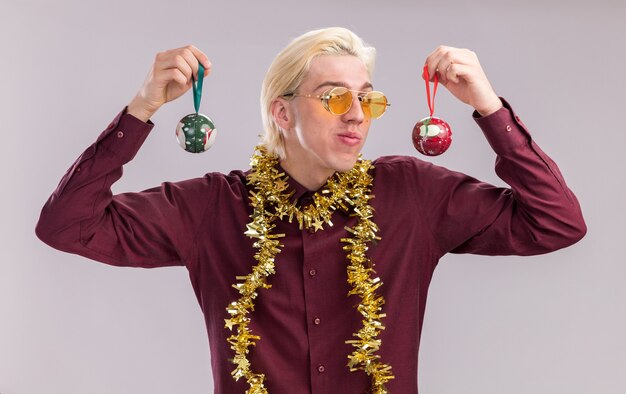 Pleased young blonde man wearing glasses with tinsel garland around neck holding christmas baubles near head  isolated on white wall