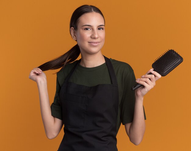 Pleased young brunette barber girl in uniform holds braid and comb on orange