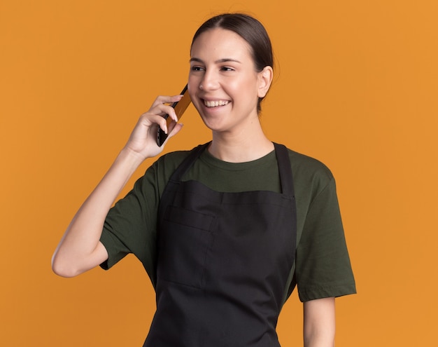 Pleased young brunette barber girl in uniform talks on phone looking at side on orange