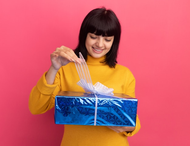 Pleased young brunette caucasian girl holds and looks at gift box isolated on pink wall with copy space