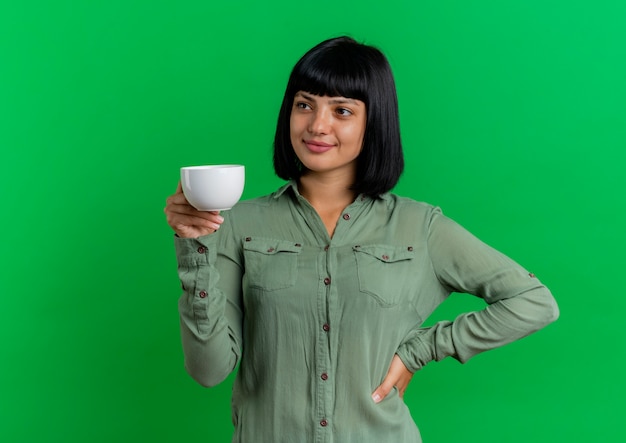 Pleased young brunette caucasian girl puts hand on waist holding cup isolated on green background with copy space