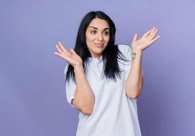 Pleased young brunette caucasian girl raises hands looking at side isolated on purple wall