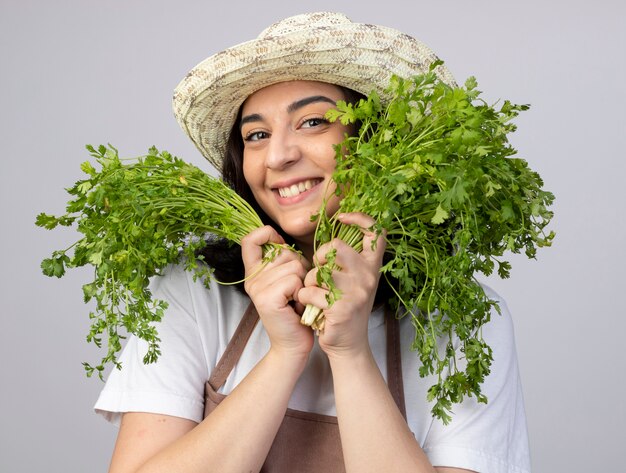 Pleased young brunette female gardener in uniform wearing gardening hat holds coriander isolated on white wall with copy space