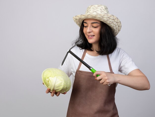 Pleased young brunette female gardener in uniform wearing gardening hat holds hoe rake and looks at cabbage isolated on white wall with copy space