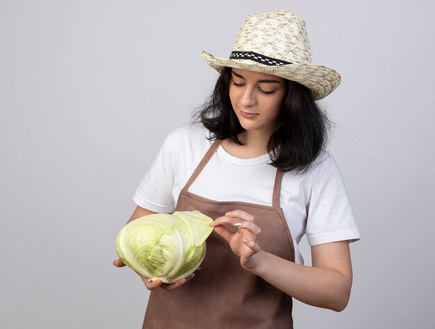 Pleased young brunette female gardener in uniform wearing gardening hat holds and looks at cabbage isolated on white wall with copy space