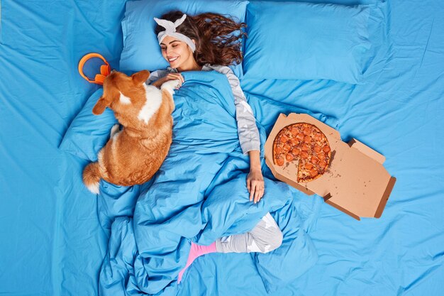 Pleased young brunette woman plays with dog dressed in comfortable pajama being lazy to get out of bed eats tasty pizza forgets about all work relaxes together with favorite pet after good sleep