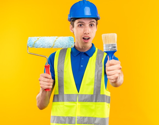 Pleased young builder man in uniform holding roller brush with paint brush 