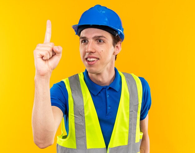 Pleased young builder man in uniform showing one isolated on yellow wall