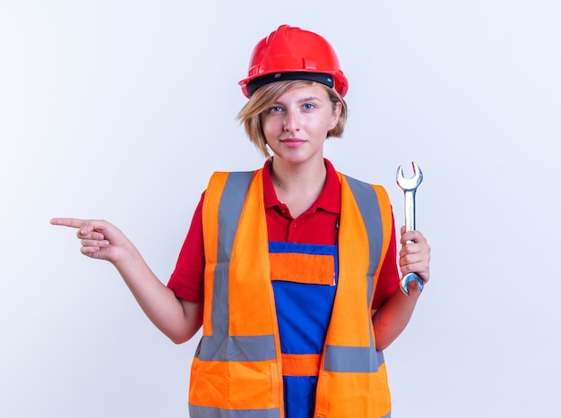 pleased young builder woman in uniform holding open-end wrench points at side isolated on white wall with copy space