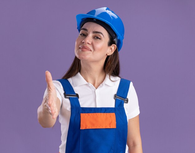 Pleased young builder woman in uniform holding out hand at camera isolated on purple wall