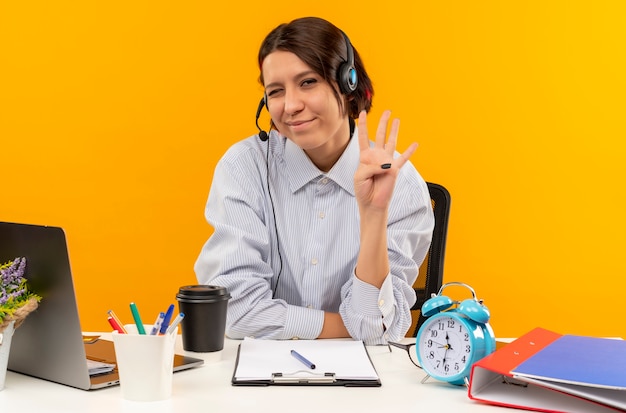 Free photo pleased young call center girl wearing headset sitting at desk winking and showing four with hand isolated on orange background