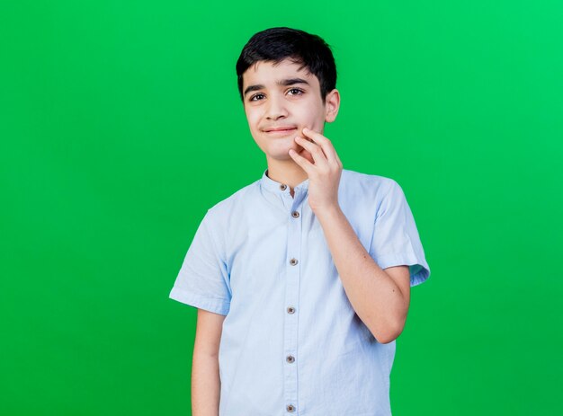 Pleased young caucasian boy  touching chin isolated on green wall with copy space