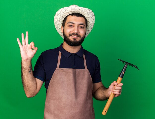 pleased young caucasian male gardener wearing gardening hat holding rake and gesturing ok sign isolated on green wall with copy space