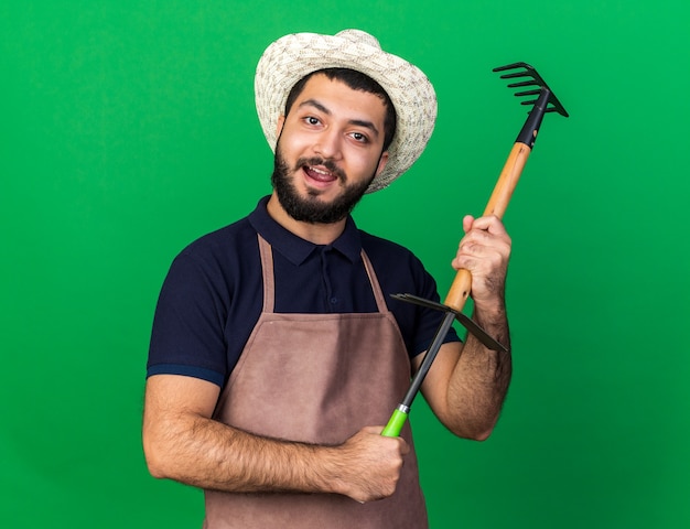 pleased young caucasian male gardener wearing gardening hat holding rake over hoe rake isolated on green wall with copy space