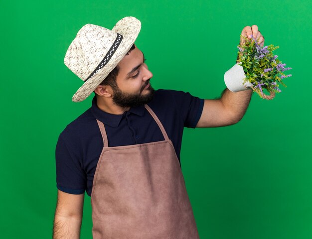 pleased young caucasian male gardener wearing gardening hat looking at flowerpot isolated on green wall with copy space