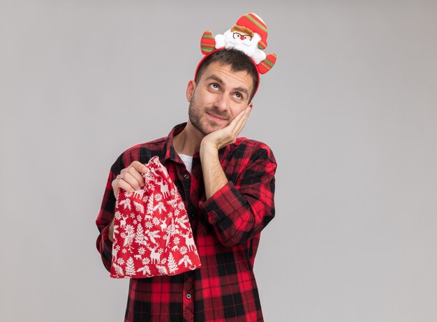 Pleased young caucasian man wearing christmas headband holding christmas sack keeping hand on face looking up isolated on white background with copy space