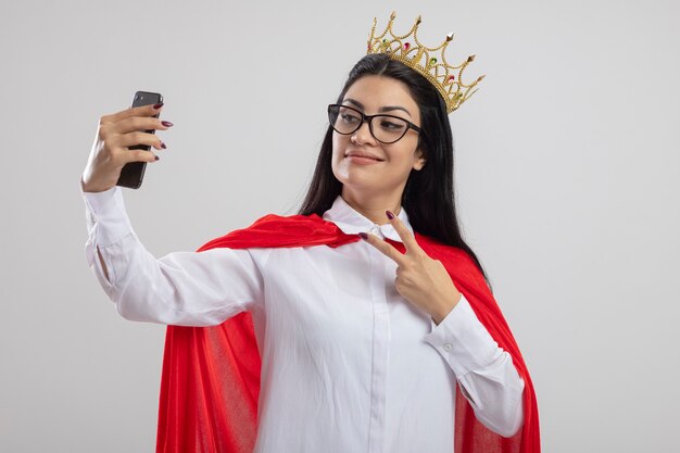 Pleased young caucasian superhero girl wearing glasses and crown doing peace sign taking selfie isolated on white background with copy space