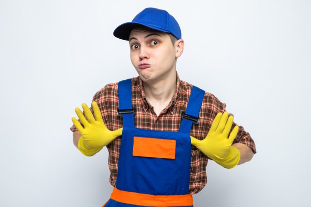 Pleased young cleaning guy wearing uniform and cap with gloves 