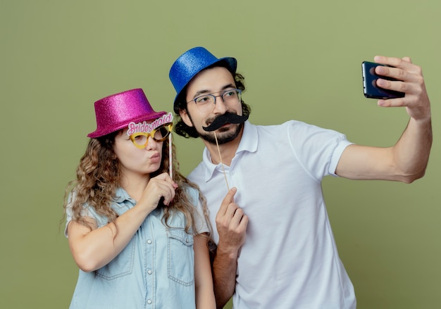 Pleased young couple wearing pink and blue hat take a selfie  girl holding masquerade eye mask on stick and guy holding fake mustache on stick 