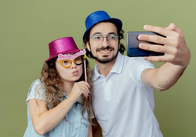 Pleased young couple wearing pink and blue hat take a selfie and girl holding masquerade eye mask on stick 