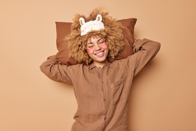 Free Photo pleased young curly woman keeps soft pillow behind head applies hydrogel patches under eyes dressed in pajama smiles happily enjoys good morning isolated over brown background time for sleeping