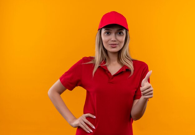 Pleased young delivery girl wearing red uniform and cap putting hand on hip her thumb up isolated on orange background